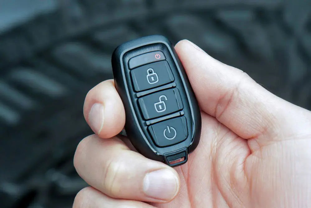 Close-up of a hand holding a black remote key fob with lock, unlock, and power buttons against a tire background