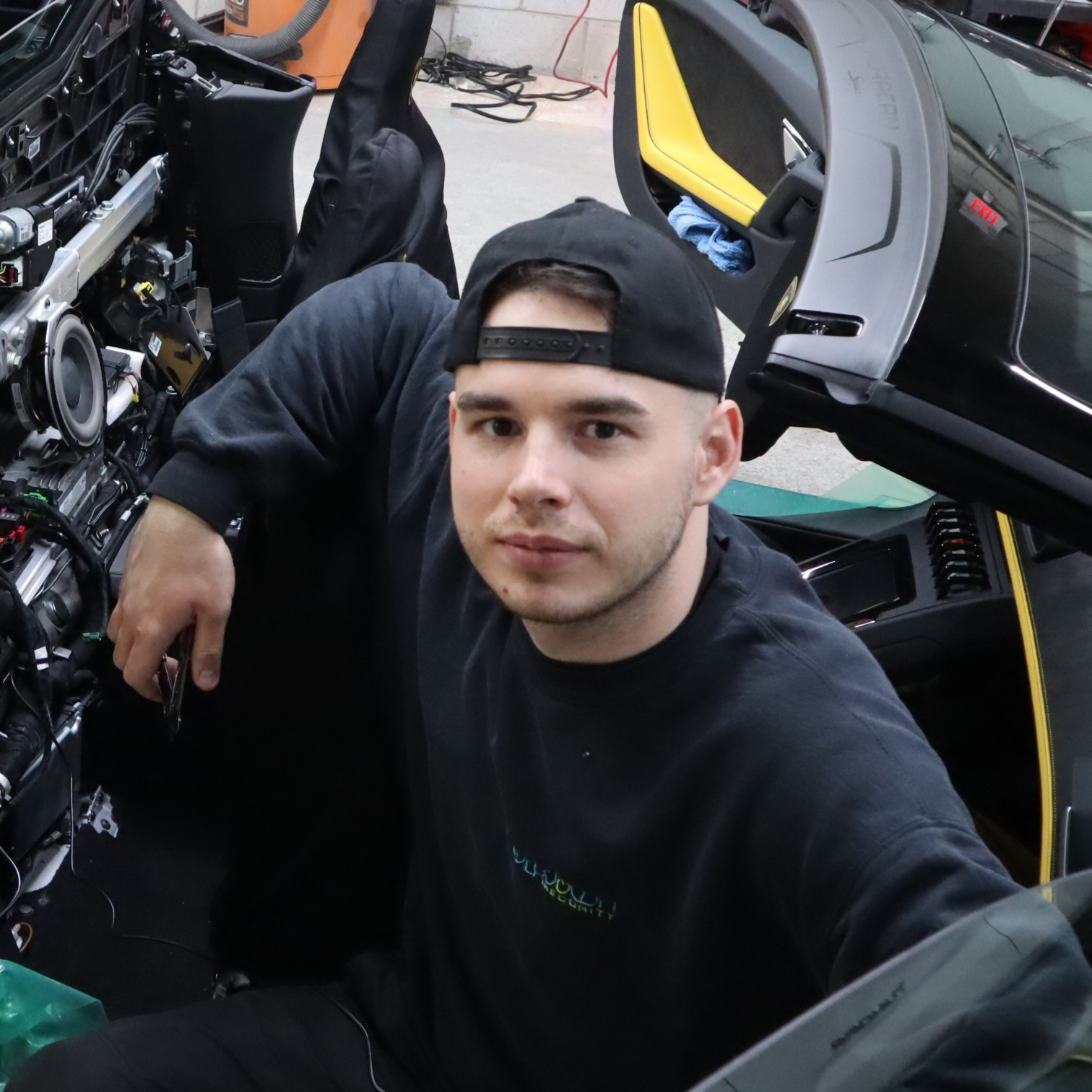 Technician wearing black cap and sweatshirt working on car interior with door open in a professional garage setting