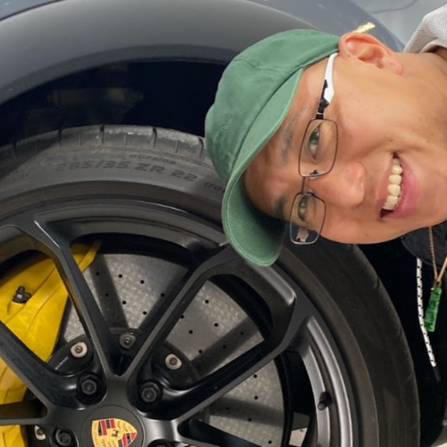 Man wearing glasses and green cap smiling next to a car wheel with yellow brake caliper and Porsche logo visible