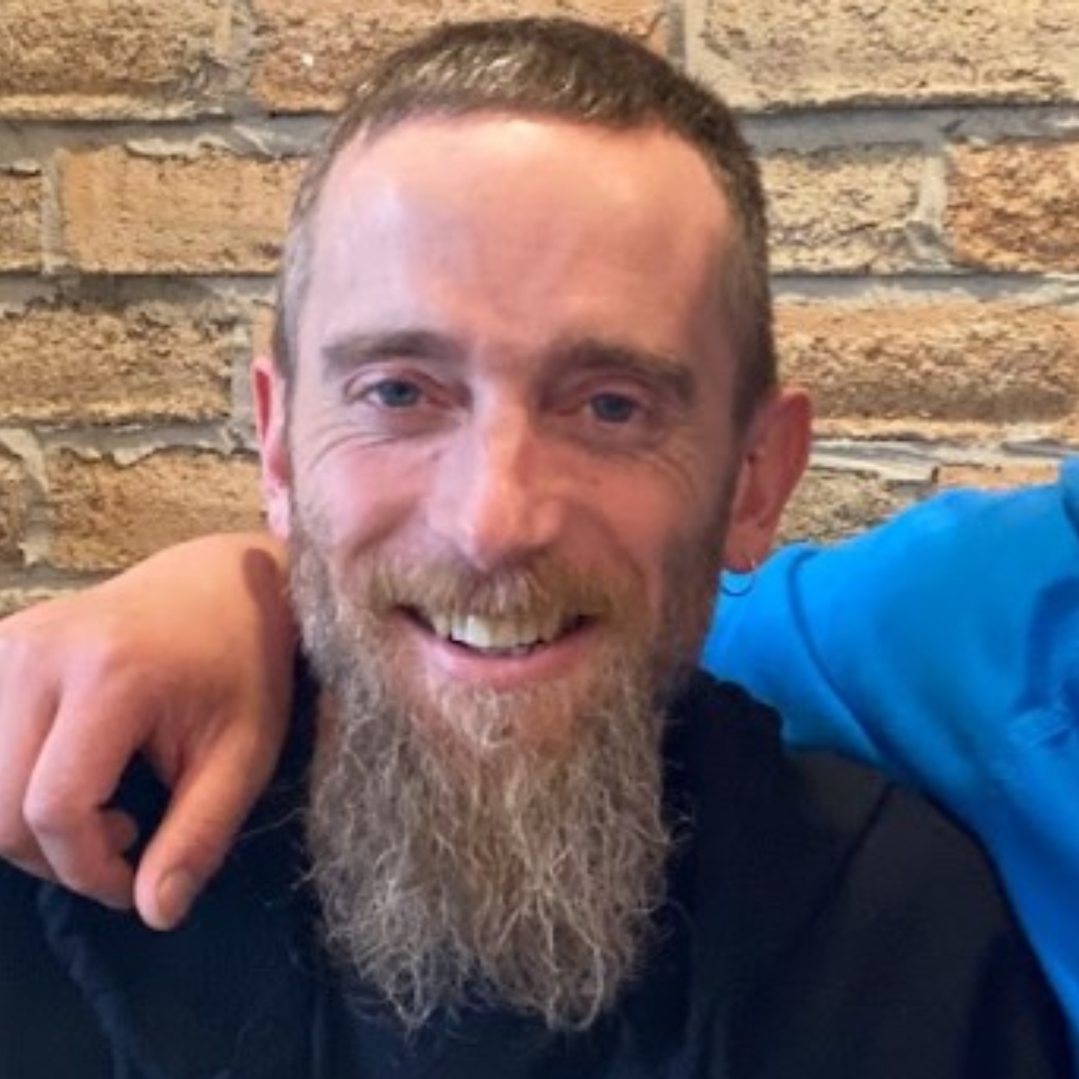 Smiling man with beard wearing a black shirt sitting indoors against a brick wall background.