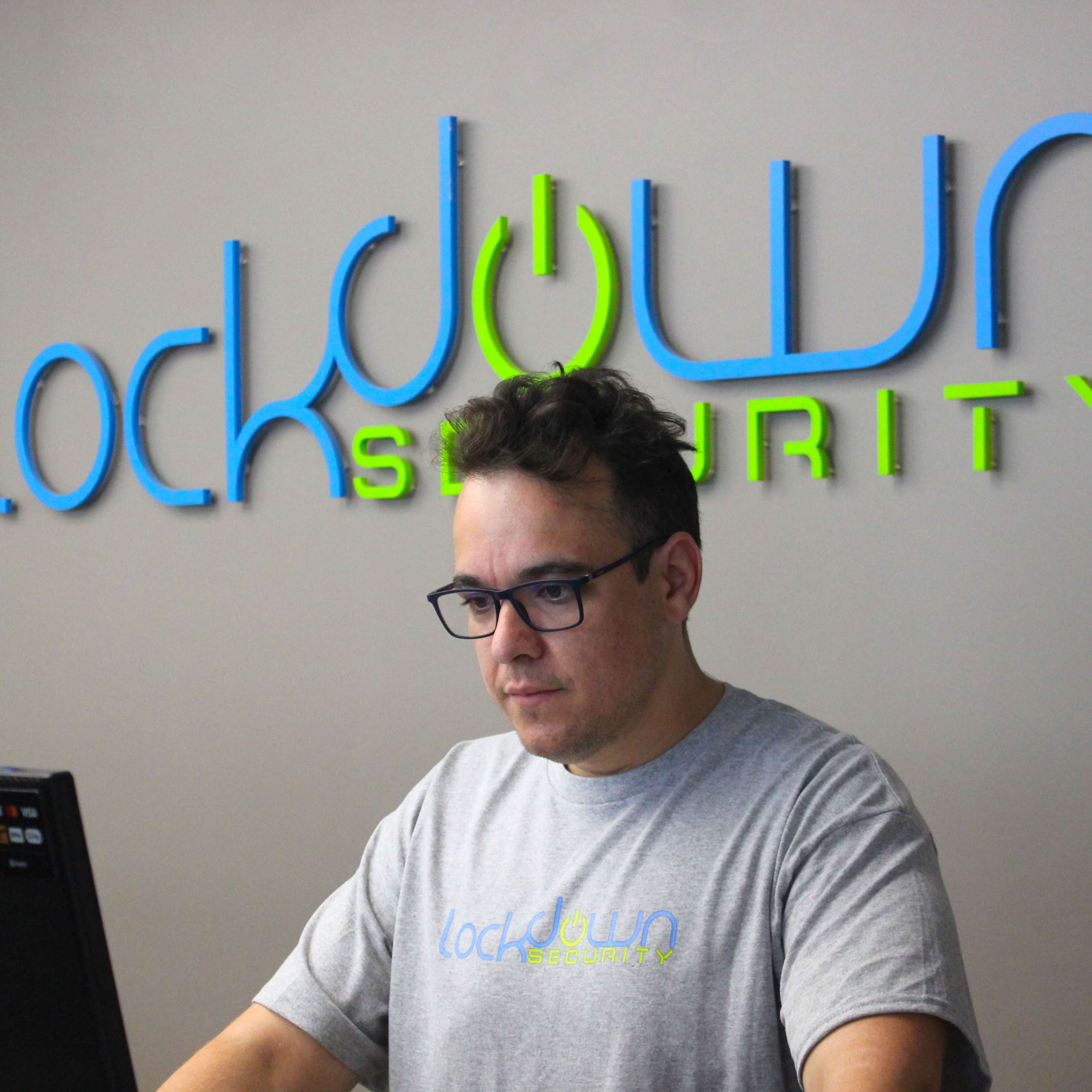 Man wearing glasses and a gray shirt working at a desk with Lockdown Security logo on wall behind him