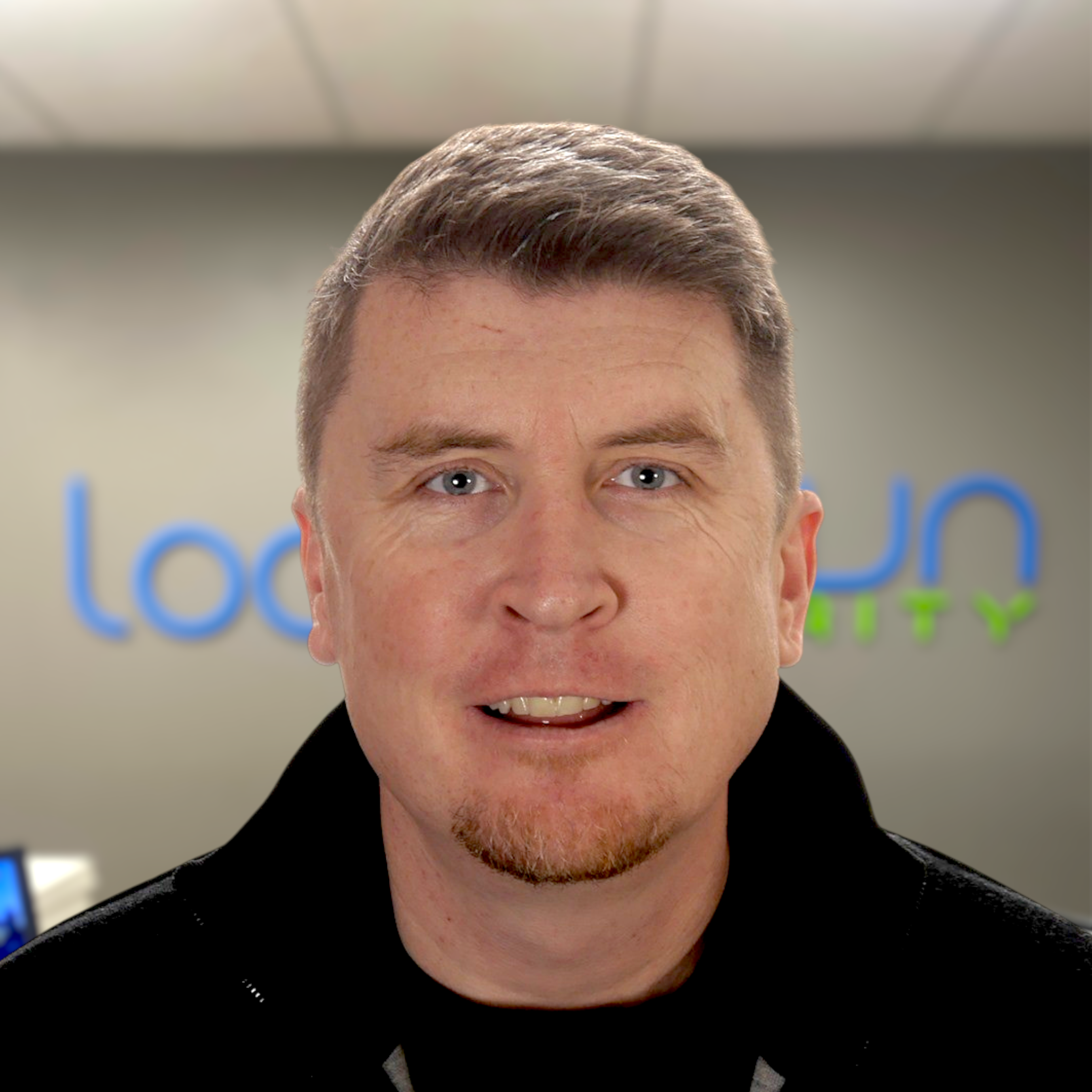 Close-up of a man with short hair and light beard smiling in an office setting with a blurred logo background.