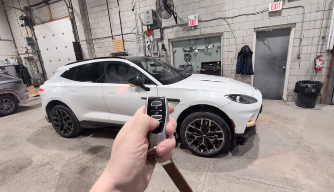 Hand holding remote key fob in front of white luxury SUV inside a vehicle security and installation garage bay.